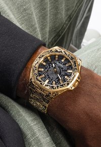 Gold-tone octagonal watch with ornate engravings, black patterned face, and luminous markers, worn on a wrist against green fabric.