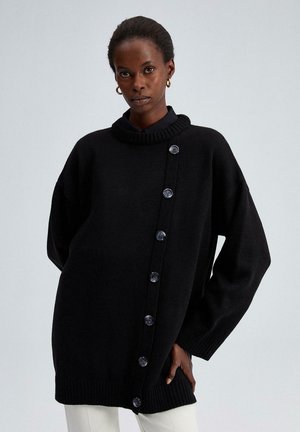 Žena oblečená v čiernom oversized pletenom svetri s diagonálne umiestnenou radou gombíkov, skombinovaná s bielymi nohavicami a zlatými kruhovými náušnicami, stojí pred jednoduchým pozadím.
