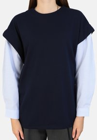 Kragenloses, dunkelblaues T-Shirt mit kurzen Ärmeln; darunter befinden sich lange Ärmel mit weißen Nadelstreifen, die einen Layer-Look und einen Kontrast in den Stoffen erzeugen.