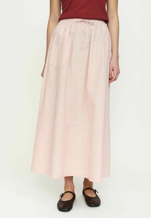 Kobieta ubrana w jasnoróżową spódnicę do kostek z wiązaniem w talii i czarne buty typu Mary Jane, stojąca na jednolitym tle.