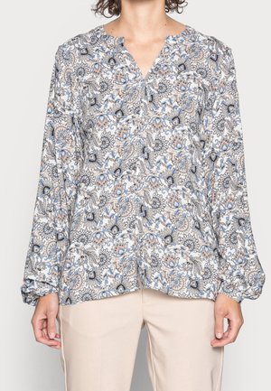 Vrouw in een blouse met lange mouwen in een blauw, beige en zwart bloemen paisley patroon, gecombineerd met lichtbeige broeken.