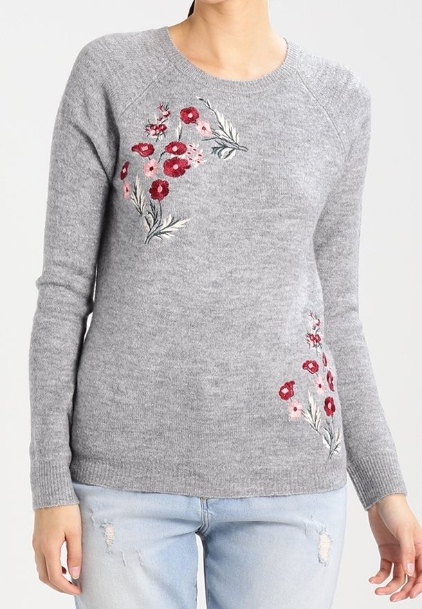Pull en maille gris avec des fleurs brodées rouges et roses sur la poitrine et le bas, porté avec un jean bleu clair déchiré.