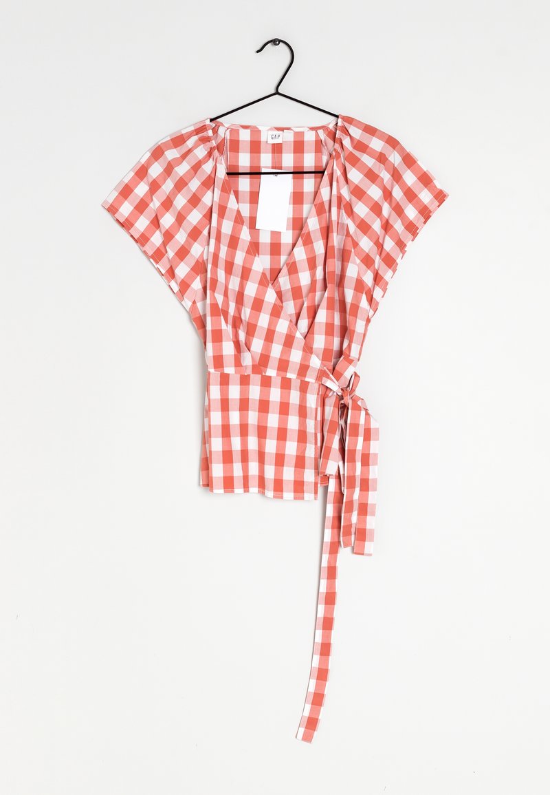Blouse portefeuille à carreaux vichy rouge et blanc à manches courtes avec un lien à la taille, suspendue sur un cintre noir sur fond blanc.