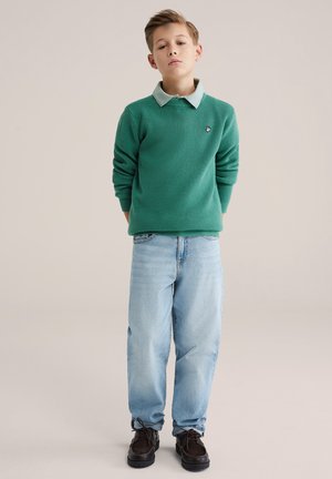 Pull vert texturé avec une chemise colletée vert clair en dessous, associé à un jean ample bleu clair et des chaussures marron foncé.