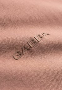 Tissu rose pâle avec une finition texturée ; logo "GABBA" embossé dans une teinte subtile assortie. Lignes épurées et éléments de design minimalistes.