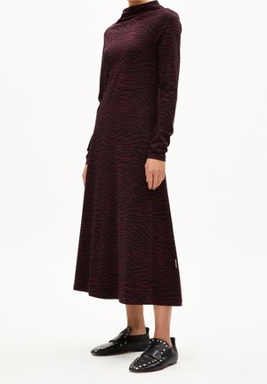 Femme portant une robe midi à manches longues rouge foncé et noire imprimée zèbre, associée à des chaussures plates en cuir noir cloutées.