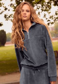 Haut zippé en velours côtelé gris avec des manches longues et un col montant. Associé à un pantalon jogger assorti avec une taille ajustable.