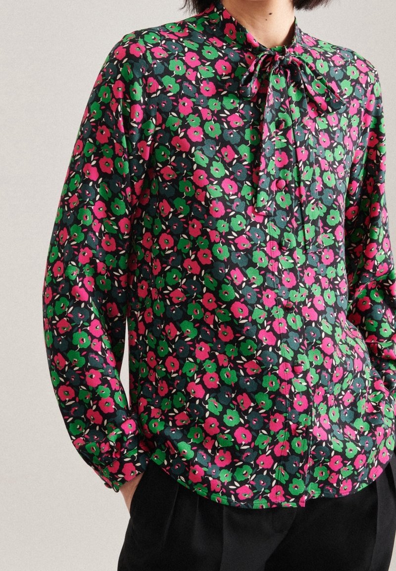 Blouse à manches longues présentant un motif floral sombre avec des fleurs roses et vertes. La texture soyeuse et le détail du col noué rehaussent le design.