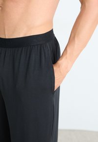 Torso masculin portant un pantalon de lounge noir Calvin Klein, avec une main dans la poche, se tenant devant un fond clair.