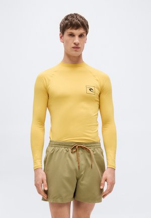 Άνδρας που φοράει μακρυμάνικη κίτρινη μπλούζα προστασίας με λογότυπο και χακί μαγιό με κορδόνι, στέκεται μπροστά από απλό φόντο.