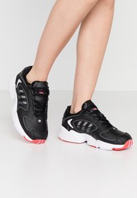 Černé tenisky s hladkým koženým svrškem, bílou mezipodešví, červenými akcenty na podrážce a pruhovaným designem po stranách. Gumová podešev.