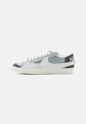 Baskets basses Nike grises et blanches avec lacets blancs, grand swoosh blanc et patch noir au talon avec texte Nike blanc.