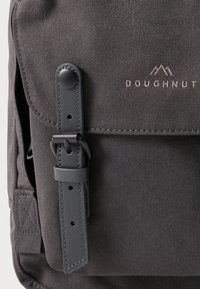 Grauer Canvas-Rucksack mit einer Frontklappe, die mit einem Knopfverschluss und einem Ledergriff ausgestattet ist. Geprägtes "DOUGHNUT"-Logo in Gold. Genähte Details.