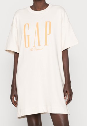 Biała sukienka oversize z krótkim rękawem z dużym, brzoskwiniowym logo "GAP The Original" na piersi, noszona przez modelkę o ciemnej karnacji, neutralne tło.