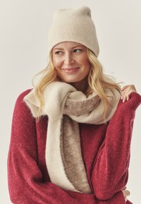 Gorro beige de punto, bufanda en crema y marrón sobre un suéter rojo. Textura suave con un diseño relajado, destacando el contraste de colores y las capas.