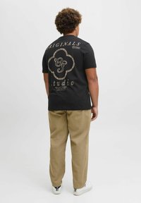 Sort kortærmet t-shirt med gyldent grafisk tekst på ryggen. Sammen med beige bukser og hvide sneakers.