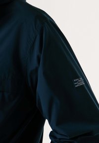 Veste imperméable bleu marine avec une texture lisse, dotée d'un logo en patch sur la manche. Vue en gros plan mettant en avant les détails de la couture de l'épaule.