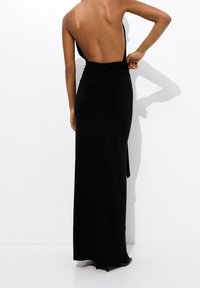 Robe longue noire dos nu en tissu doux, dotée d'une ceinture froncée et d'une fente latérale pour faciliter les mouvements.