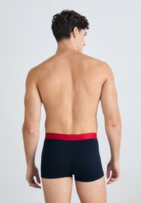 Navyfarbene Boxershorts mit rotem Gummizug, hergestellt aus einem glatten Stoff. Das Design zeichnet sich durch eine enganliegende Form aus, die die Taille und Beine betont.