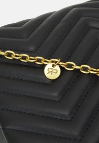 Czarna skórzana torebka z pikowanym wzorem w jodełkę, ozdobiona złotym łańcuszkiem i okrągłą zawieszką z logo przymocowaną do łańcuszka.