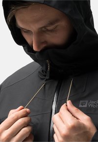 Schwarze Kapuzenjacke mit verstellbaren Kordeln, die eine glatte Textur und einen reflektierenden Reißverschluss aufweist, mit einem Branding im Brustbereich.