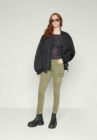 Černá bomber bunda, šedé tričko, olivově zelené cargo kalhoty a černé kotníkové boty. Modelka stojí s uvolněnýma rukama, sluneční brýle rámují její tvář.