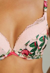 Reggiseno floreale rosa con bordo in pizzo, coppe imbottite e spalline regolabili, caratterizzato da un motivo floreale rosso e verde su uno sfondo rosa chiaro.