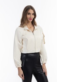 Giacca crema tagliata, con zip frontale, colletto rialzato e maniche testurizzate. Pantaloni neri aderenti contrastano con il materiale morbido della giacca.