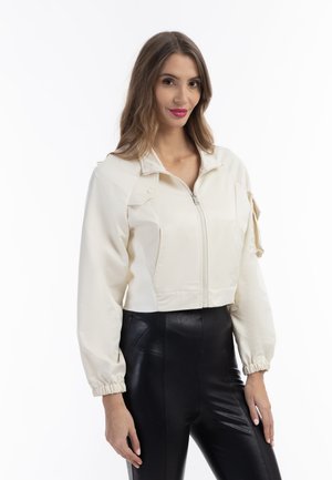 Giacca crema tagliata, con zip frontale, colletto rialzato e maniche testurizzate. Pantaloni neri aderenti contrastano con il materiale morbido della giacca.