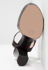 Chaussure à talon haut en cuir noir avec un bout rond, un dos ouvert et une sangle réglable. Semelle texturée avec la taille 37 moulée au bas.