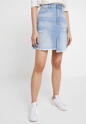 Jupe en jean bleu clair, avec une fente à l'avant, un ourlet effiloché et des patches déchirés. Portez avec des baskets blanches.