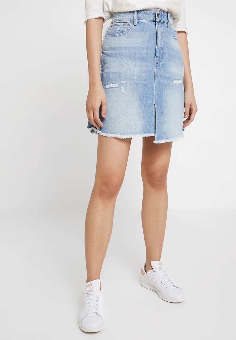 Jeanskjol i ljusblått, med en frontslits, fransad fåll och slitna lappar. Buren med vita sneakers.