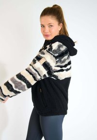 Felpa in pile con fondo nero e un motivo a righe in beige, grigio e bianco nella parte superiore. Include una tasca con zip e cappuccio.