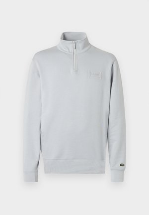Sweatshirt gris clair à col demi-zippé, poignets côtelés et logo Lacoste texturé sur la poitrine, fabriqué en tissu doux.