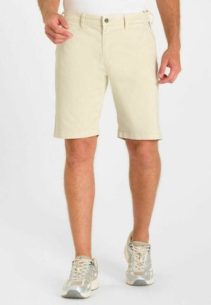 Pantaloncini beige fino al ginocchio indossati da una persona in piedi con scarpe sportive di tonalità chiara su uno sfondo semplice.