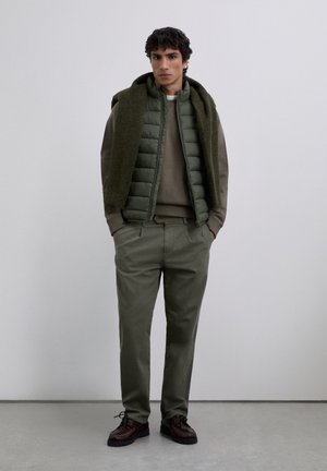 Mann in olivgrünem gepolstertem Westen über braunem Pullover, olivfarbener Hose, dunkelbraunen Schuhen und grünem Schal, der über die Schultern gelegt ist, steht drinnen.