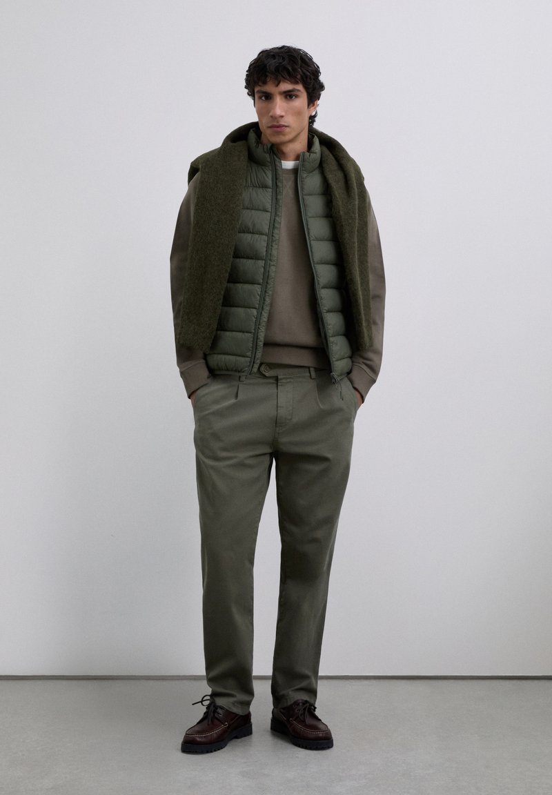 Man wearing olive green padded vest over brown sweater, olive trousers, dark brown shoes, and green scarf draped over shoulders, standing indoors.