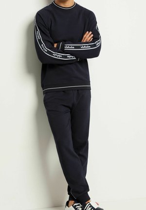 Osoba ubrana w czarny zestaw Athleisure z białym napisem „vitula” na rękawach, czarne spodnie i czarno-białe sneakersy, oparta o białą ścianę.