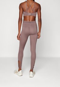 Bra sportivo marrone con spalline incrociate, abbinato a leggings a vita alta con ritagli sulle caviglie. Scarpe da ginnastica bianche completano l'outfit.