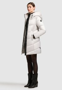 Puffjakke i skinnende hvid, med hætte og sidelommer. Har et diagonalt quiltet design og sort lynlås, parret med elegante sorte støvler.