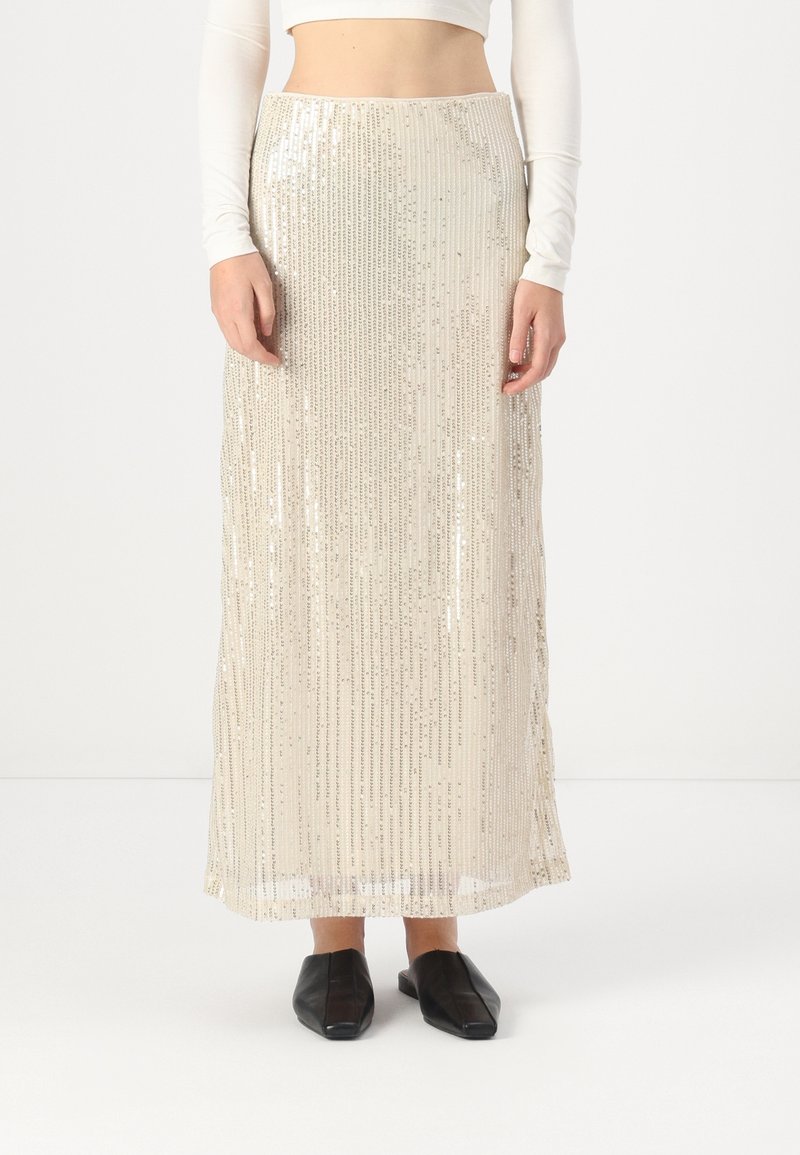 Jupe longue beige taille haute avec des sequins argentés verticaux, portée avec un crop top blanc à manches longues et des chaussures plates noires à bout carré.