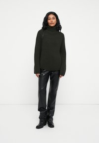 Dunkelgrüner, gerippter Rollkragenpullover, oversized Schnitt, kombiniert mit schwarzen, hochtaillierten Kunstlederhosen und schwarzen Stiefeletten.