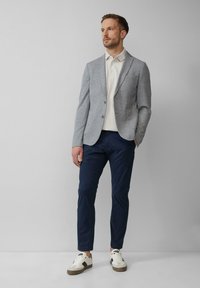 Blazer gris texturé avec revers crantés sur un polo blanc, associé à un pantalon bleu marine et des baskets blanches avec des accents marron.