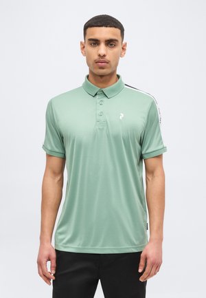 Homme portant un polo à manches courtes vert clair avec une bande noire et blanche sur l'épaule, debout devant un fond clair uni.