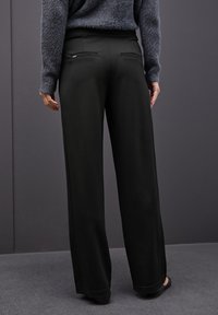 Schwarzweite Hosen mit glattem Stoff, bestehend aus einem hohen Taillenbund und zwei hinteren Taschen, von hinten vor einem neutralen Hintergrund gezeigt.