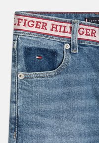 Džínové kalhoty s světlým modrým odstínem, s bílým pasem, na kterém je červený text "HILFIGER" a malou logem nášivkou poblíž kapsy.