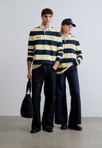 Μπλούζες με μακριά μανίκια ριγέ σε κίτρινο και ναυτικό μπλε, συνδυασμένες με σκούρο τζιν παντελόνι. Τα αξεσουάρ περιλαμβάνουν ένα μαύρο καπέλο και μια μαύρη τσάντα ώμου.