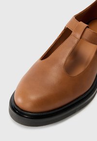 Chaussure en cuir marron avec un bout rond, design en T, semelle en caoutchouc noire et couture visible le long des bords. Texture lisse.