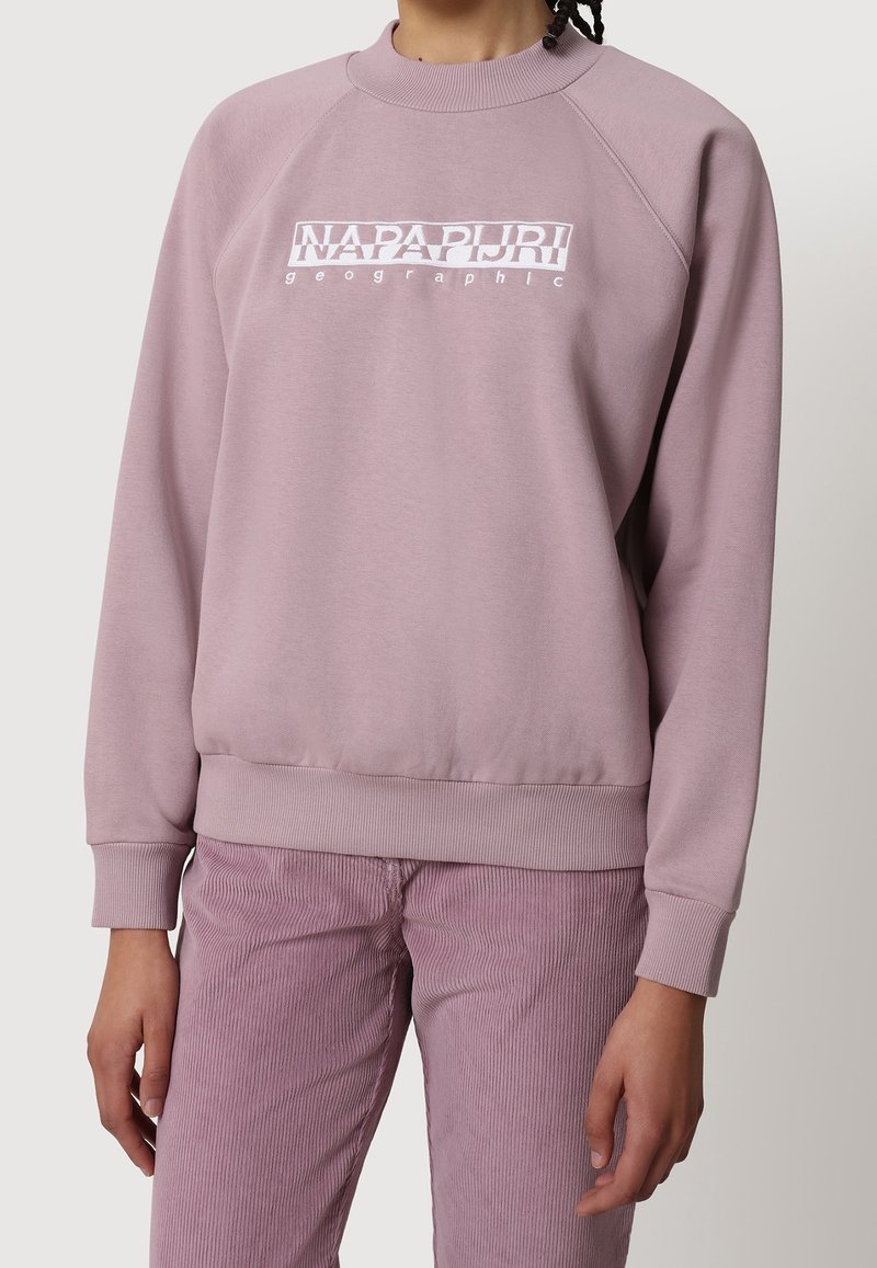 Personne portant un sweat-shirt mauve avec le logo « NAPAPIJRI geographic » et un pantalon en velours côtelé mauve assorti, sur un fond clair uni.