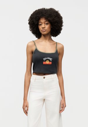 Giovane donna con capelli ricci che indossa un top nero corto con una grafica e pantaloni bianchi a vita alta, in piedi davanti a uno sfondo semplice.